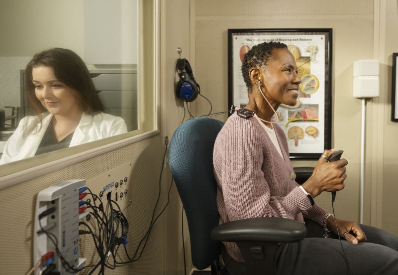 A woman sits in hearing test booth and has her hearing tests, while an audiologist reads the report.
