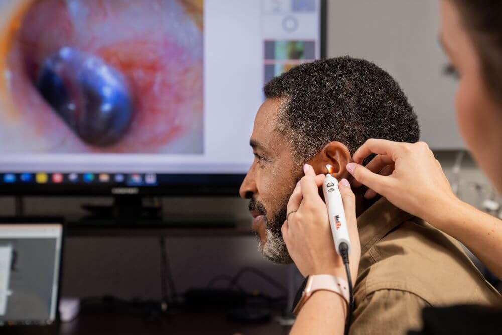 An audiologist performs a tympanometer test on a patient. The patient's eardrum is visualized on a TV screen in the background. 