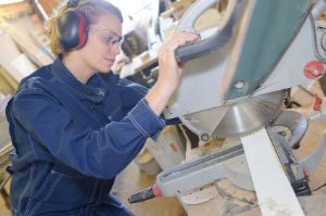 A woman operates a saw wearing ear and eye protection.
