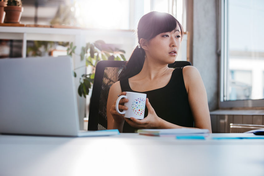 A woman sits at a work desk holding a mug of coffee.